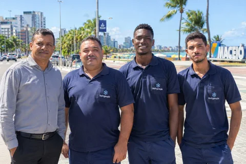 Equipe da Refrigeração Maceió reunida na orla da cidade, com o mar e o letreiro de Maceió ao fundo